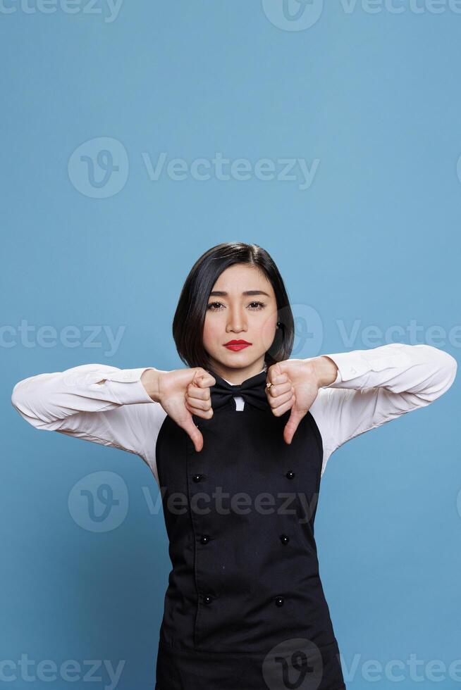 Restaurant woman employee showing thumbs down gesture, symbolizing experience discontent and dissatisfaction portrait. Asian waitress displaying disapproval concept and looking at camera photo