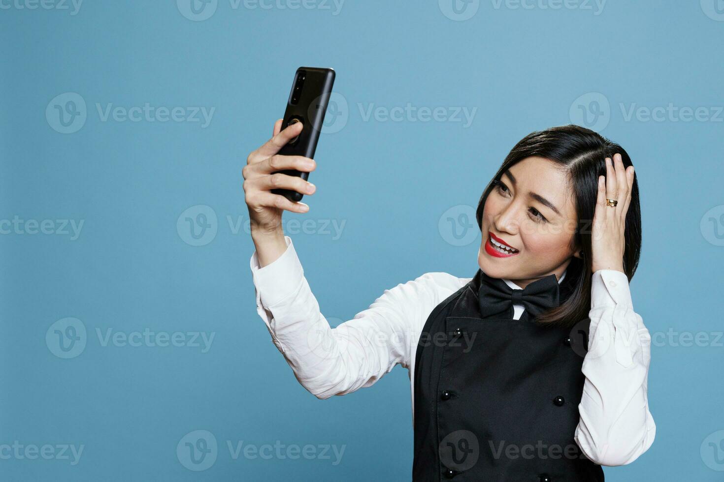 Smiling young asian waitress looking at smartphone front camera and fixing hairstyle. Cheerful woman receptionist with joyful face expression taking selfie on mobile phone photo