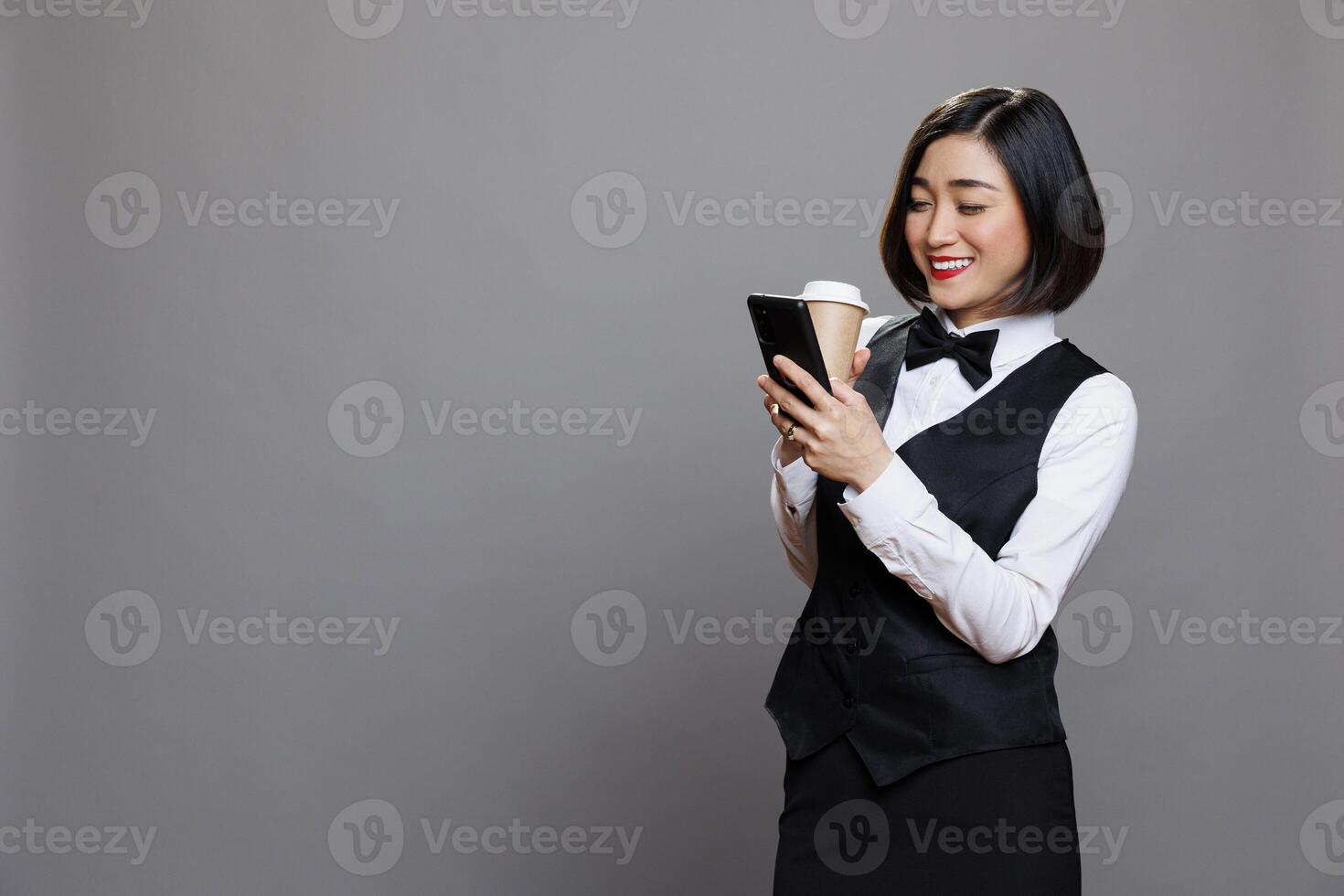 Smiling asian woman wearing receptionist uniform texting and scrolling social media while drinking coffee to go. Restaurant cheerful waitress enjoying tea in paper mug and using mobile phone photo