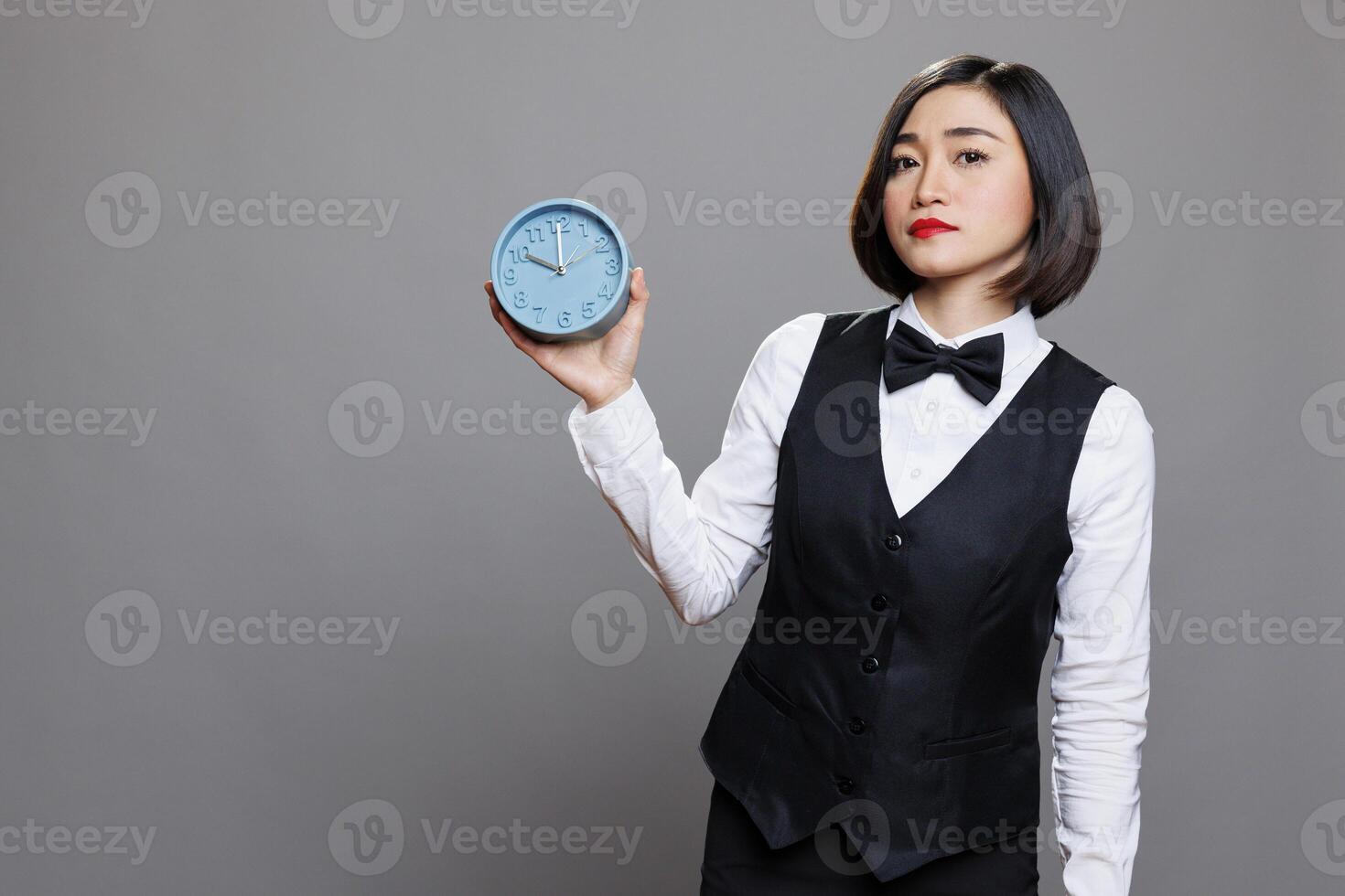 Hotel woman receptionist holding retro alarm clock and looking at camera with serious facial expression. Young asian waitress wearing uniform showing time management concept portrait photo