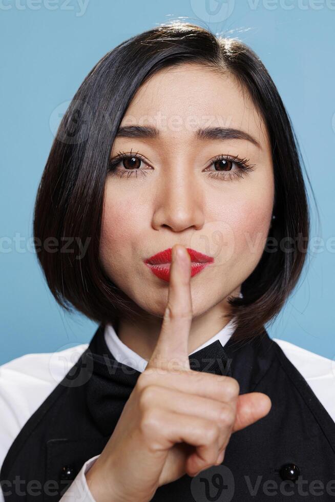 Serious asian woman in waitress uniform holding forefinger on lips while showing tsss gesture and looking at camera. Young receptionist asking for silence closeup portrait photo