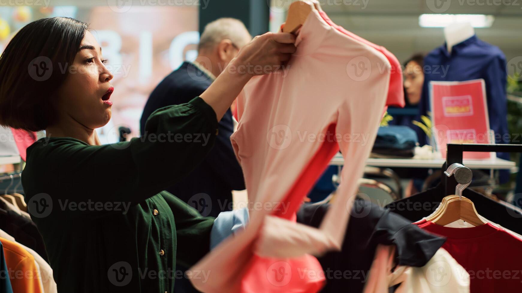 Asian woman pick clothes in a hurry to benefit from black friday seasonal sales and special offers. Shopping obsessed client grabbing many clearance items, bargain hunting rush. Handheld shot. photo