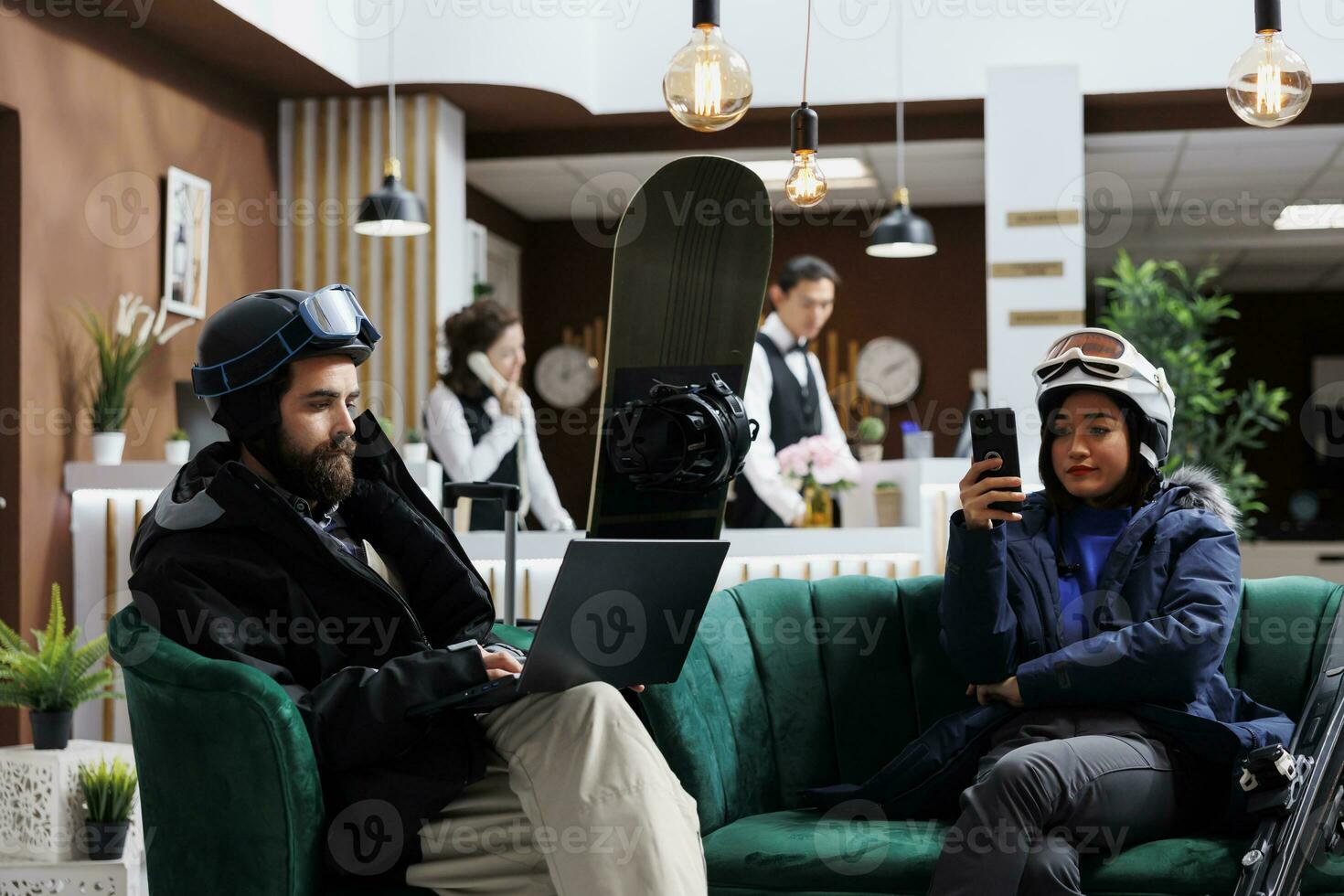 Boyfriend using laptop and girlfriend with mobile phone enjoying digital communication. Wintersports enthusiasts in hotel lobby for ski and snowboarding fun waiting with their smart devices. photo