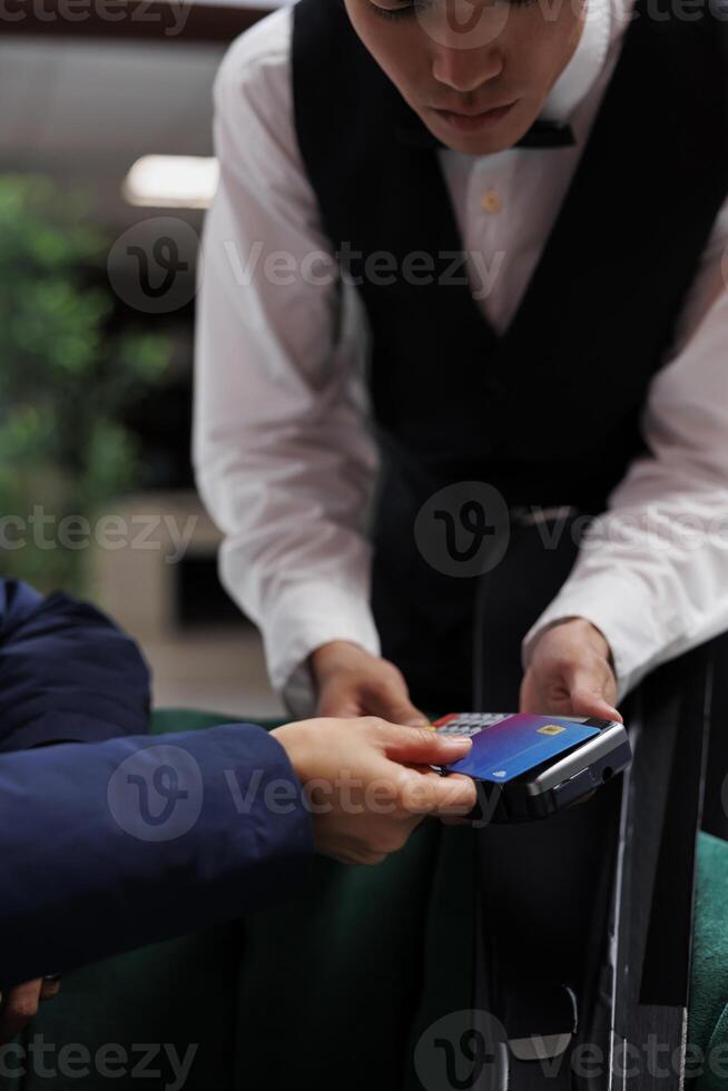 Close up of male concierge grasping pos machine and facing customer paying with debit card in hotel lobby. In lounge area of the ski resort a detailed view of credit card used for cashless payments. photo