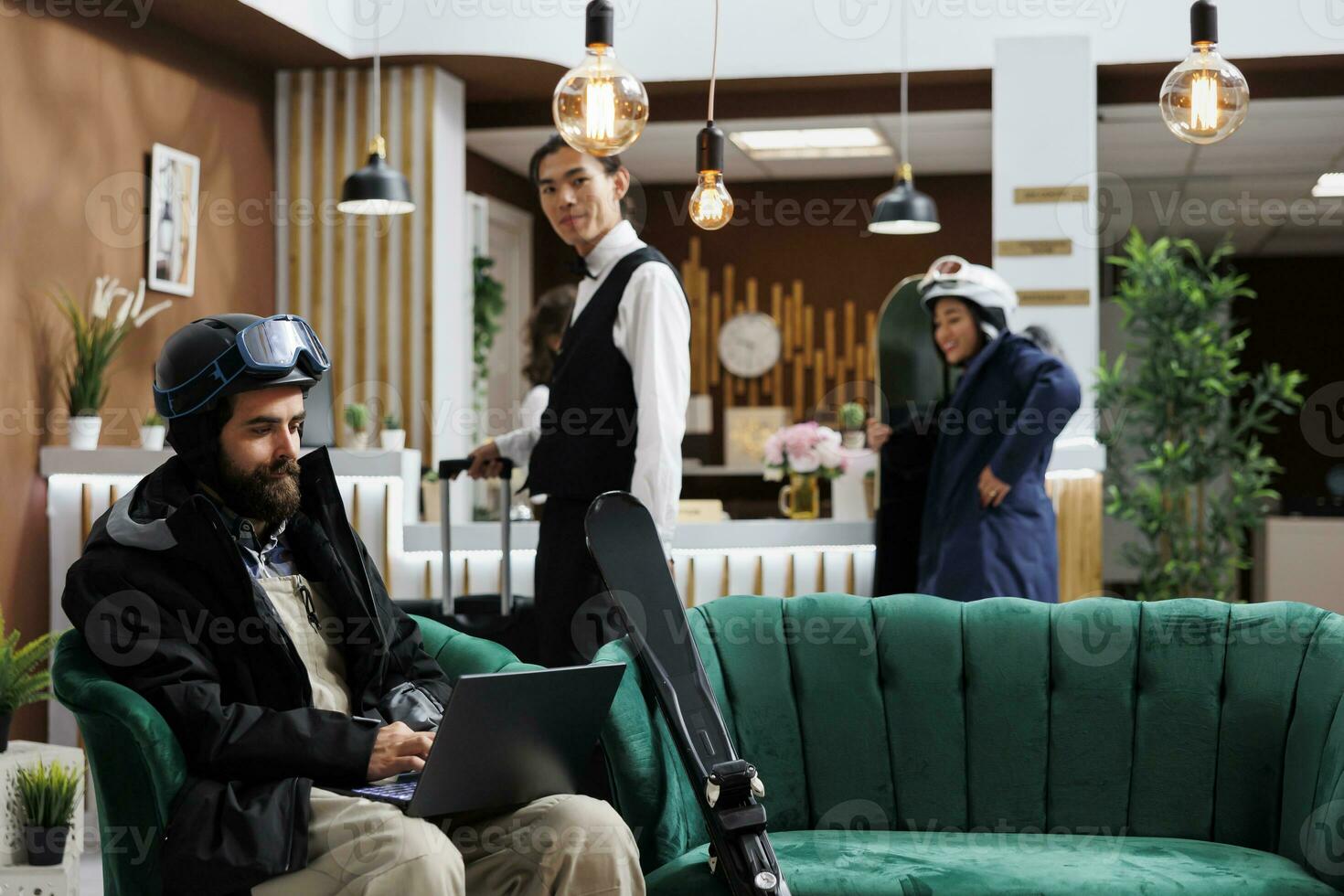 Caucasian man utilizing laptop to book activities for winter vacation at ski resort. Male guest dressed in snow clothing using minicomputer in lounge area while employee takes suitcase to hotel room. photo