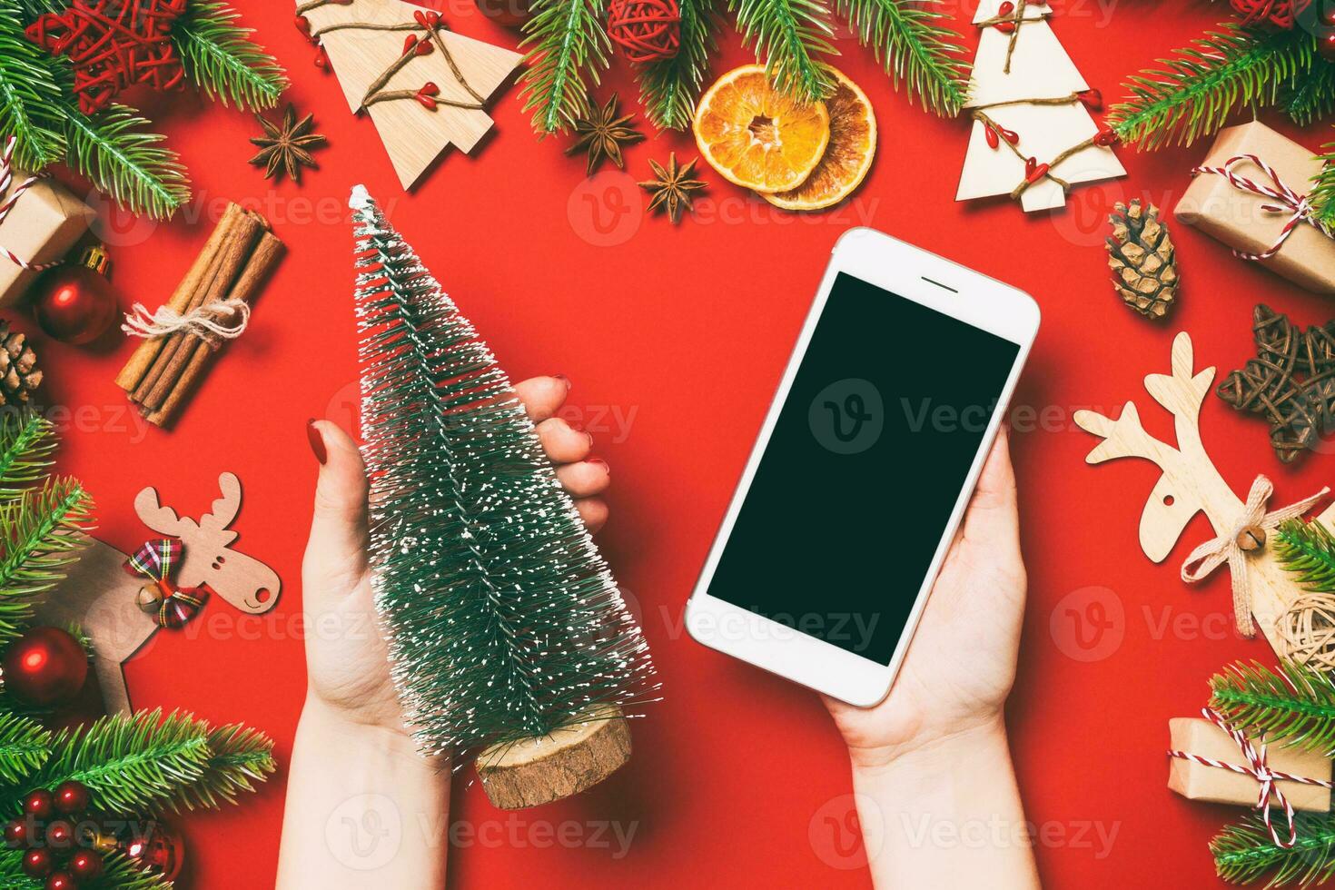 Top view of female hands holding phone in one hand and christmas tree in another hand on red background. New Year holiday concept. Mockup photo