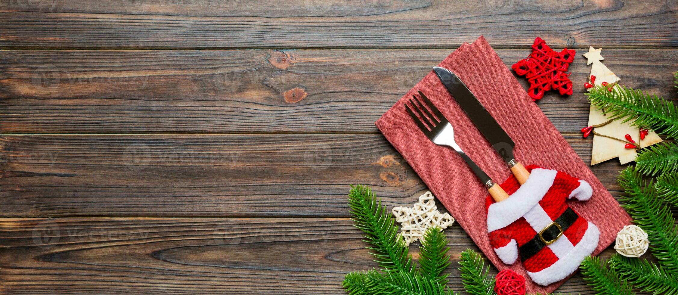 Top view of fork and knife put in santa clothes on napkin with christmas decorations and new year tree on wooden background. Banner holiday and festive concept with copy space photo