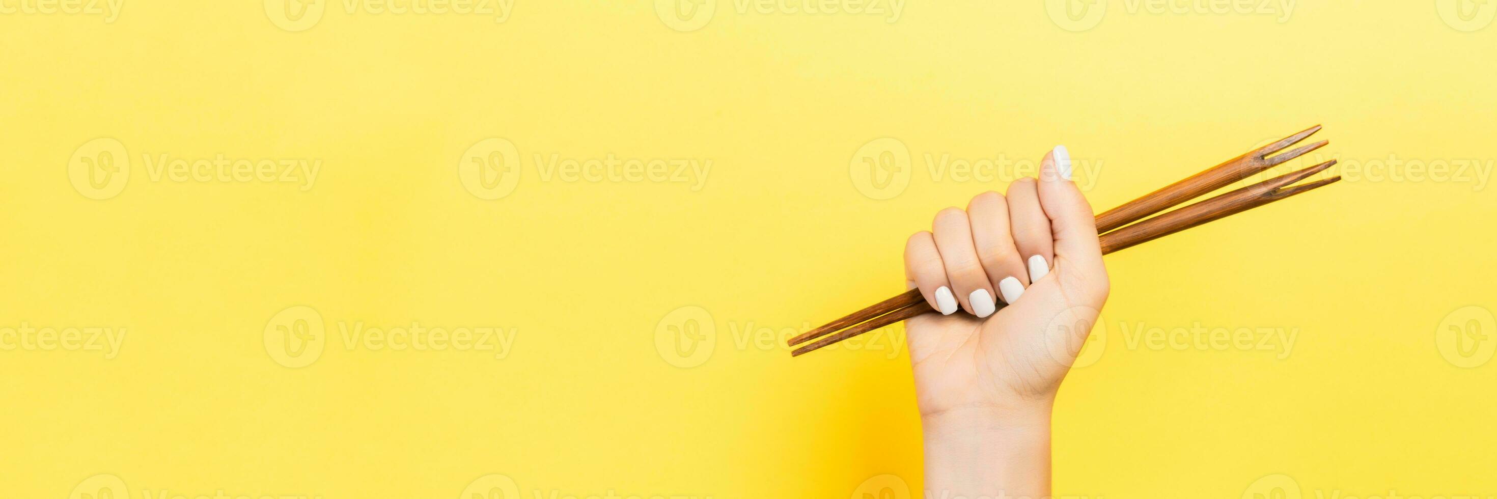 Cropped image of female hand holding chopsticks in fist on yellow background. Asian food concept with copy space photo