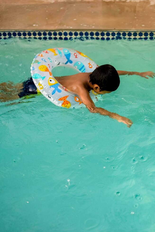 Happy Indian boy swimming in a pool, Kid wearing swimming costume along with air tube during hot summer vacations, Children boy in big swimming pool. photo