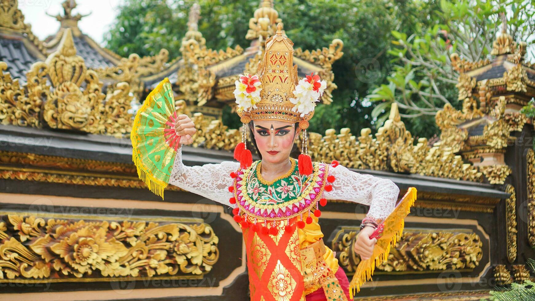 girl wearing Balinese traditional dress with a dancing gesture on Balinese temple background with hand-held fan, crown, jewelry, and gold ornament accessories. Balinese dancer woman portrait photo