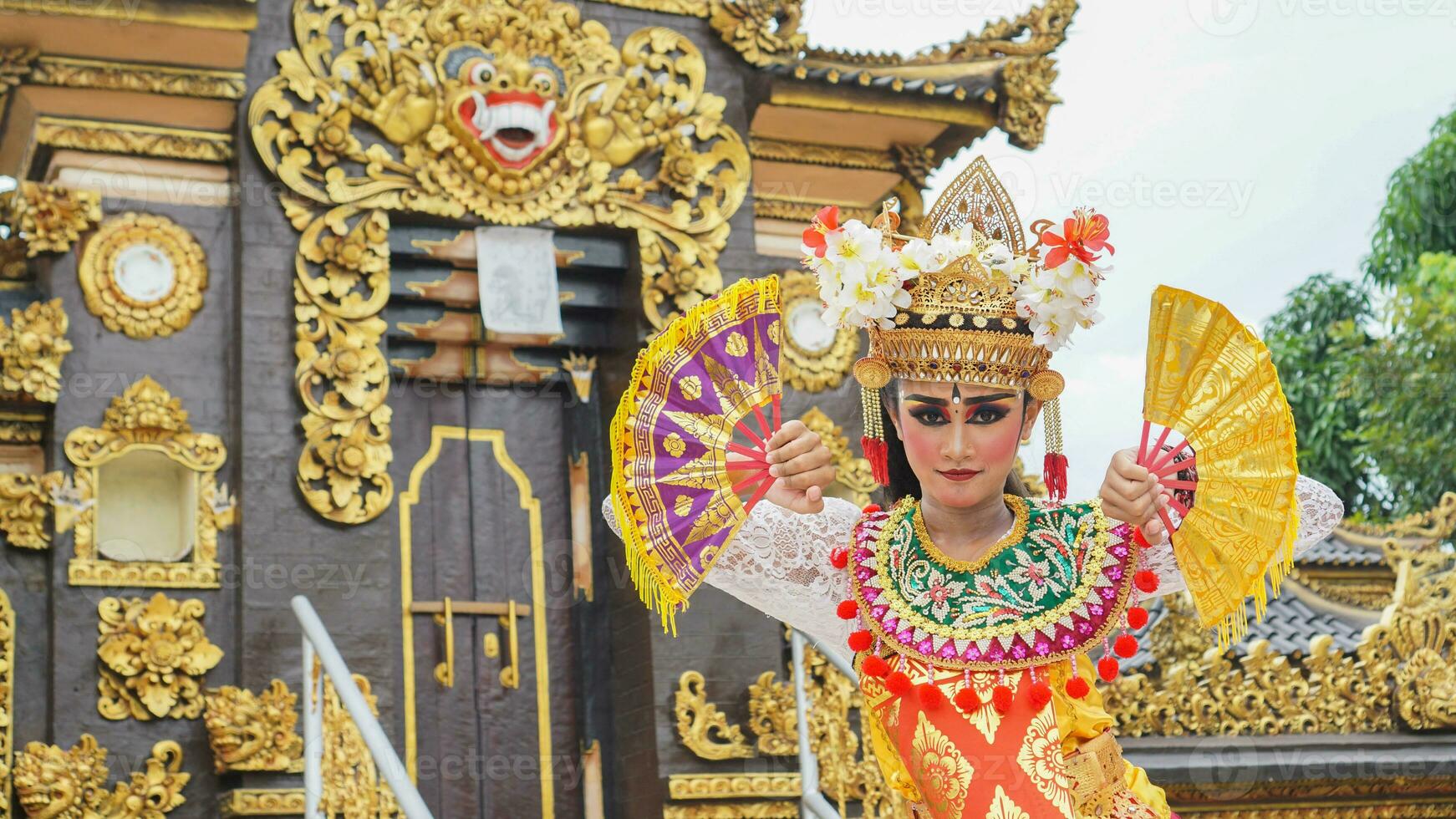 girl wearing Balinese traditional dress with a dancing gesture on Balinese temple background with hand-held fan, crown, jewelry, and gold ornament accessories. Balinese dancer woman portrait photo