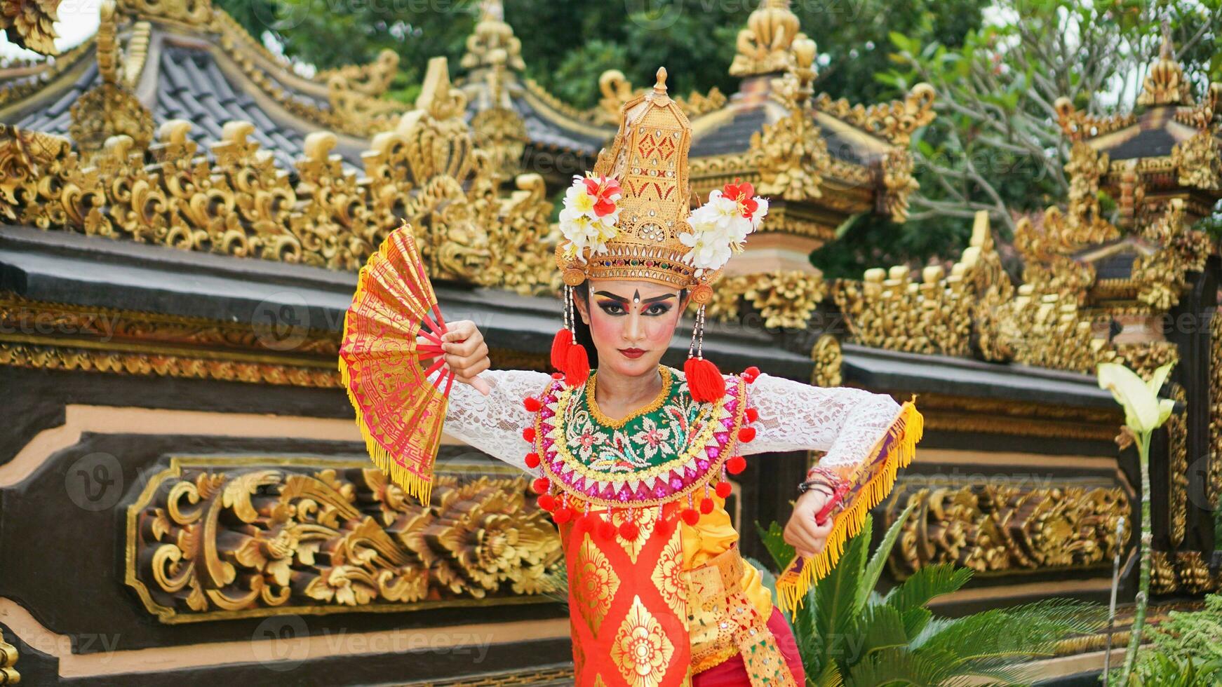 girl wearing Balinese traditional dress with a dancing gesture on Balinese temple background with hand-held fan, crown, jewelry, and gold ornament accessories. Balinese dancer woman portrait photo