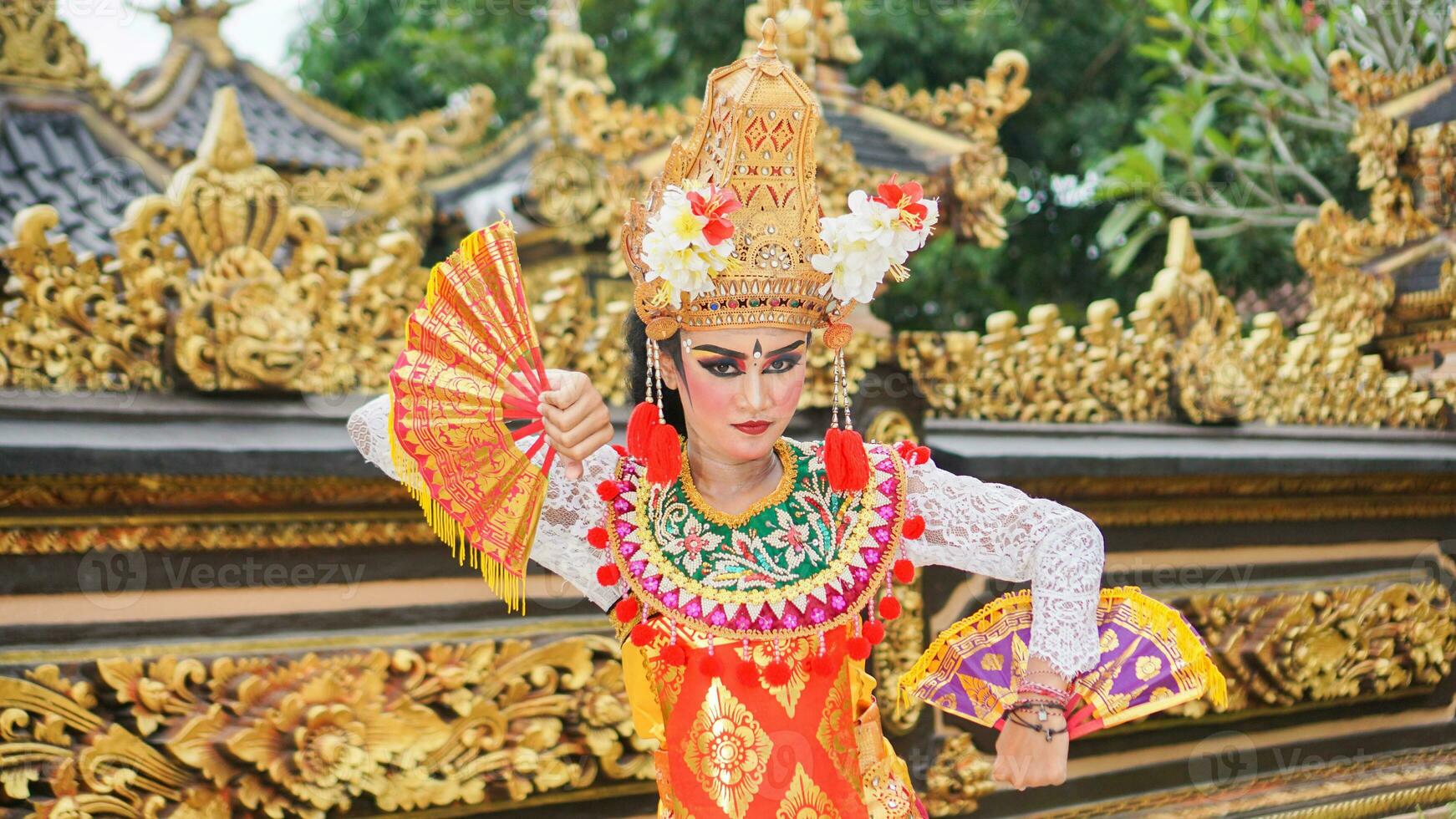 girl wearing Balinese traditional dress with a dancing gesture on Balinese temple background with hand-held fan, crown, jewelry, and gold ornament accessories. Balinese dancer woman portrait photo