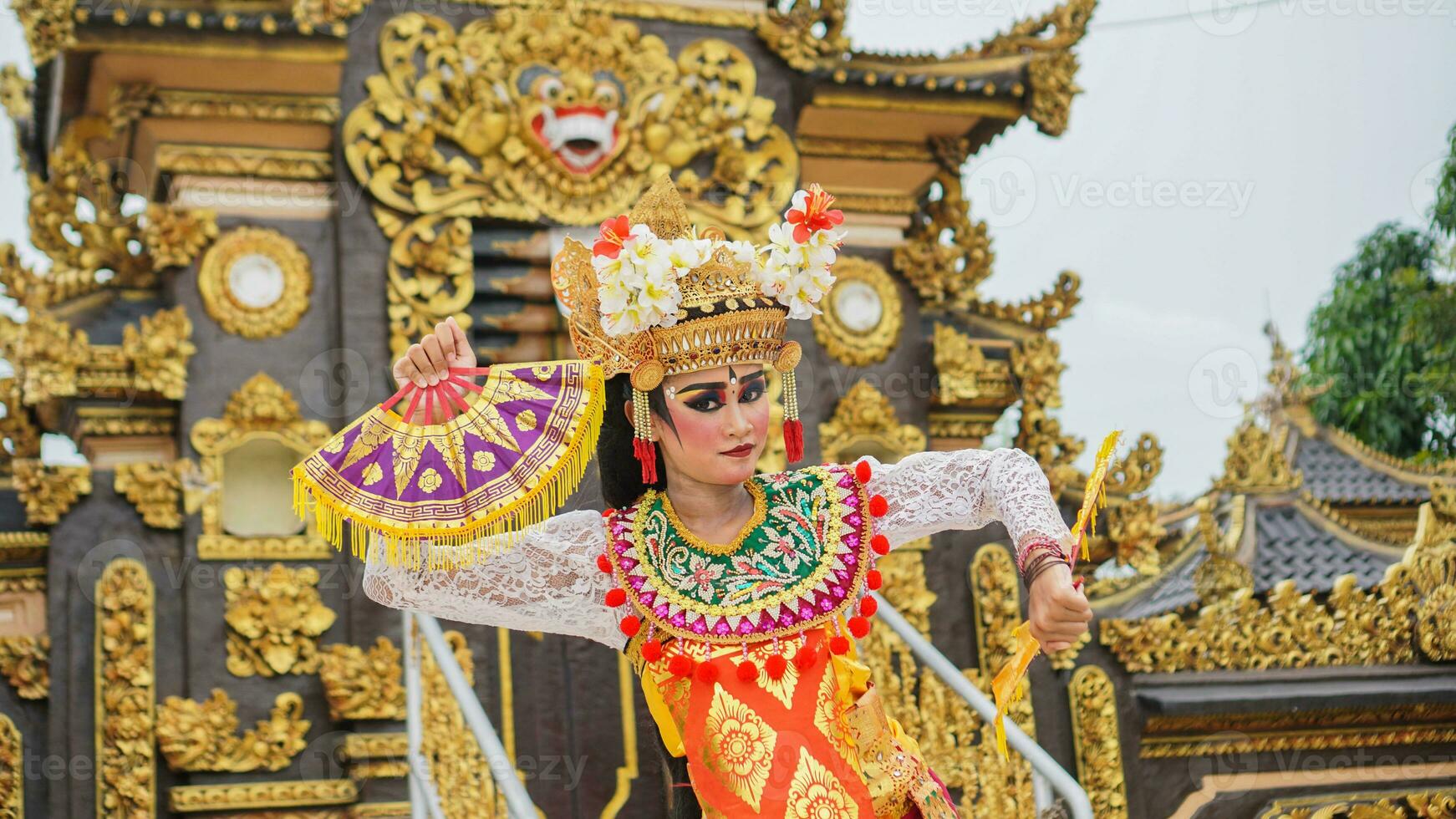 girl wearing Balinese traditional dress with a dancing gesture on Balinese temple background with hand-held fan, crown, jewelry, and gold ornament accessories. Balinese dancer woman portrait photo