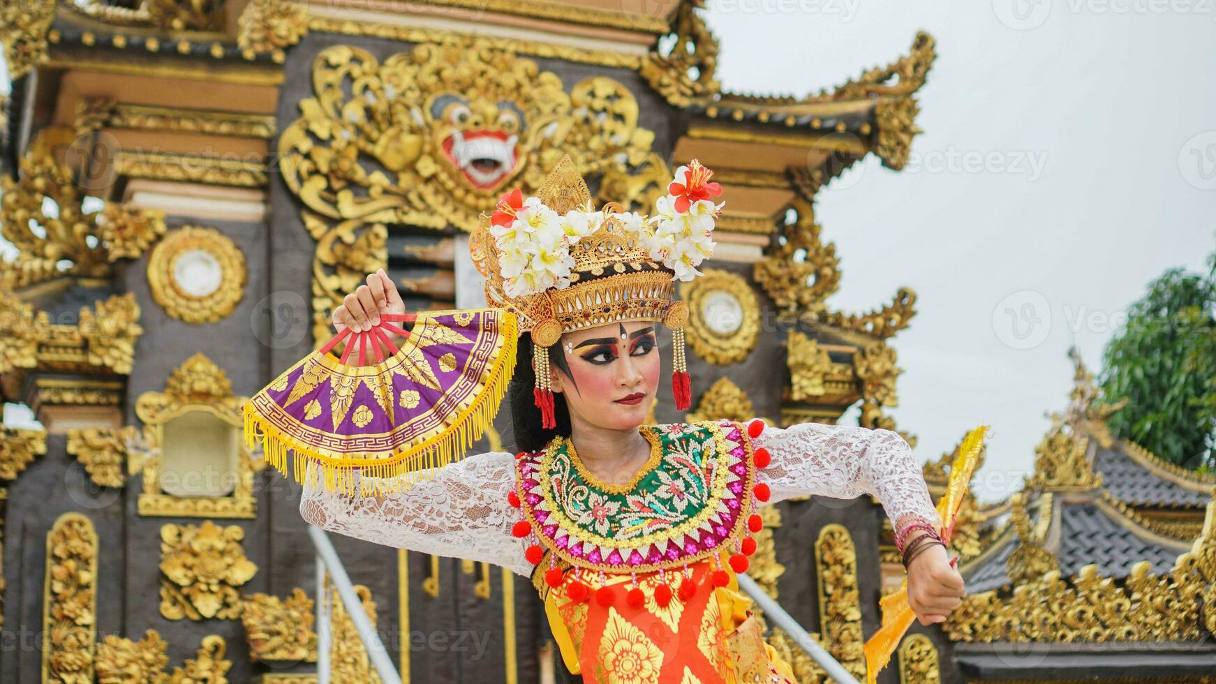 girl wearing Balinese traditional dress with a dancing gesture on Balinese temple background with hand-held fan, crown, jewelry, and gold ornament accessories. Balinese dancer woman portrait photo