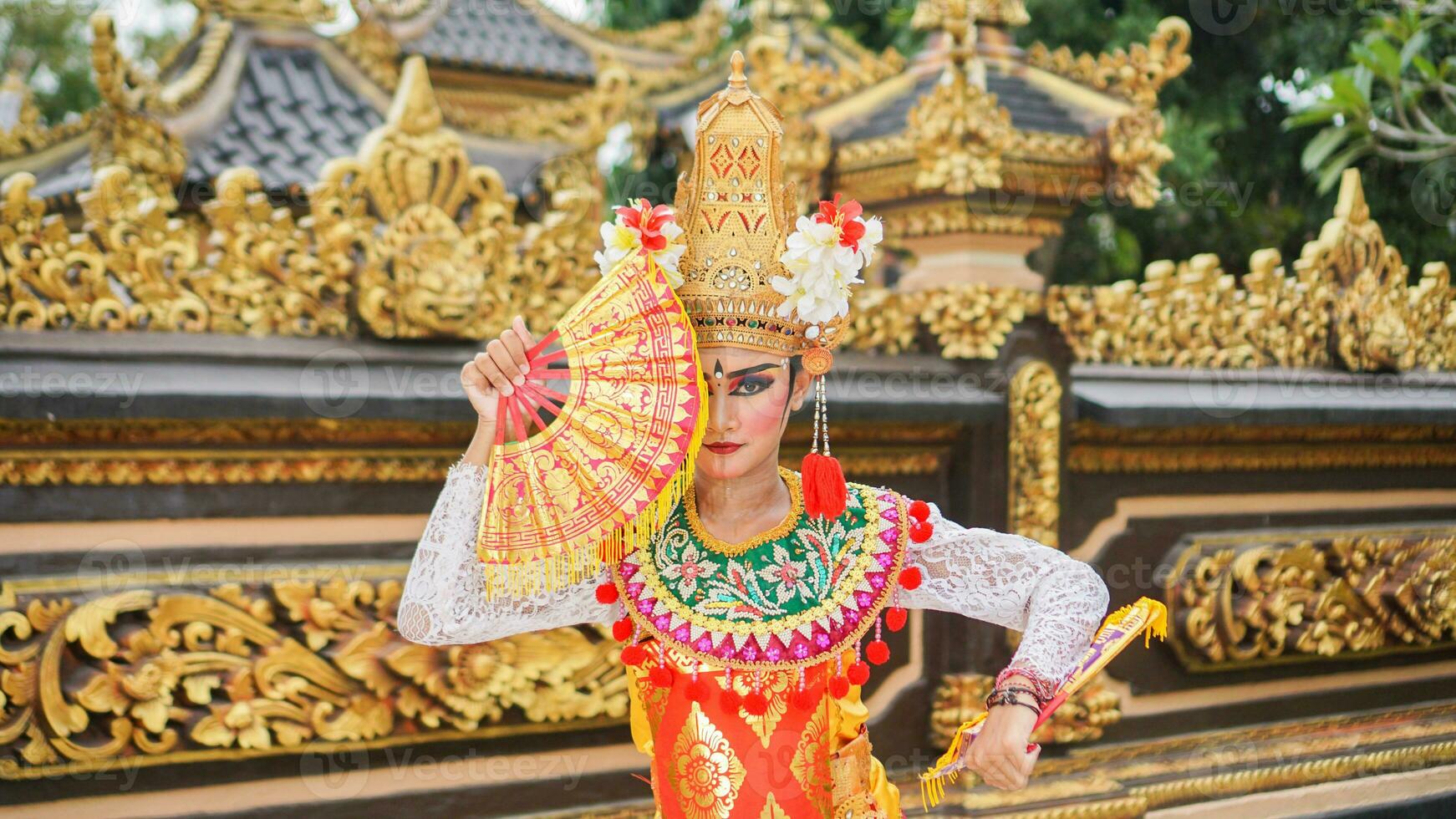 girl wearing Balinese traditional dress with a dancing gesture on Balinese temple background with hand-held fan, crown, jewelry, and gold ornament accessories. Balinese dancer woman portrait photo
