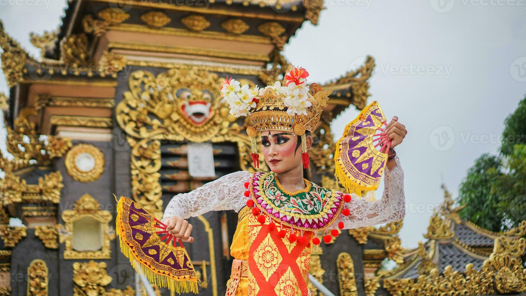 girl wearing Balinese traditional dress with a dancing gesture on Balinese temple background with hand-held fan, crown, jewelry, and gold ornament accessories. Balinese dancer woman portrait photo