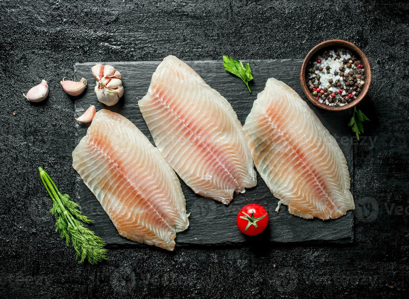 Fish fillet on black stone Board with dill, tomatoes and spices in bowl. photo