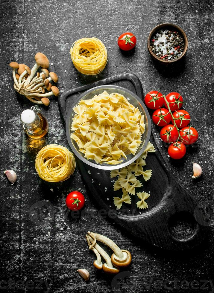 Raw farfalle and tagliatelle paste in a bowl on a cutting Board with mushrooms, tomatoes and spices. photo