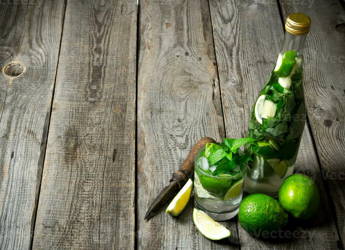 Cocktail in glass and bottle , also mint and limes on a wooden table . Free space for text. photo