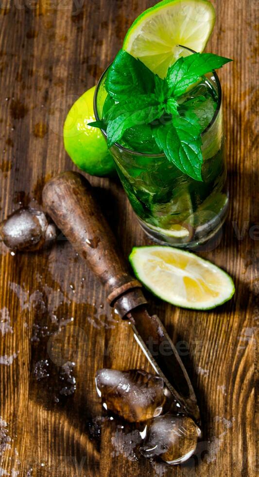 Glass of cool cocktail , ice, knife and slices of lime on a wooden table. photo