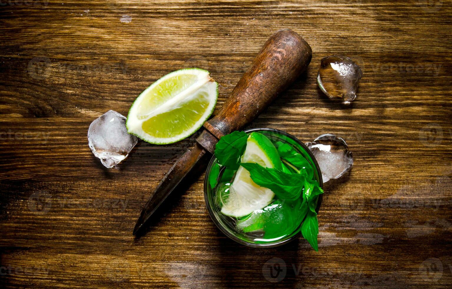 Glass of cool cocktail , ice, knife and slices of lime on a wooden table. photo