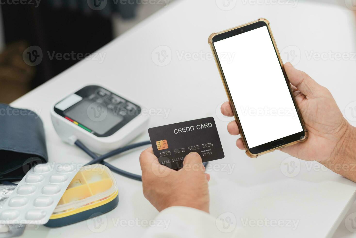 Telemedicine concept, a person holding a credit card and a smart phone on a desk with medical equipment items - blank white screen with clipping path. photo