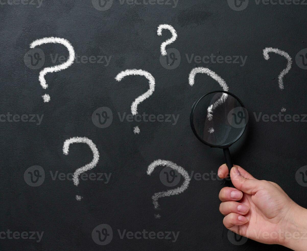 A woman's hand holds a magnifying glass on the background of a black chalk board with question marks photo