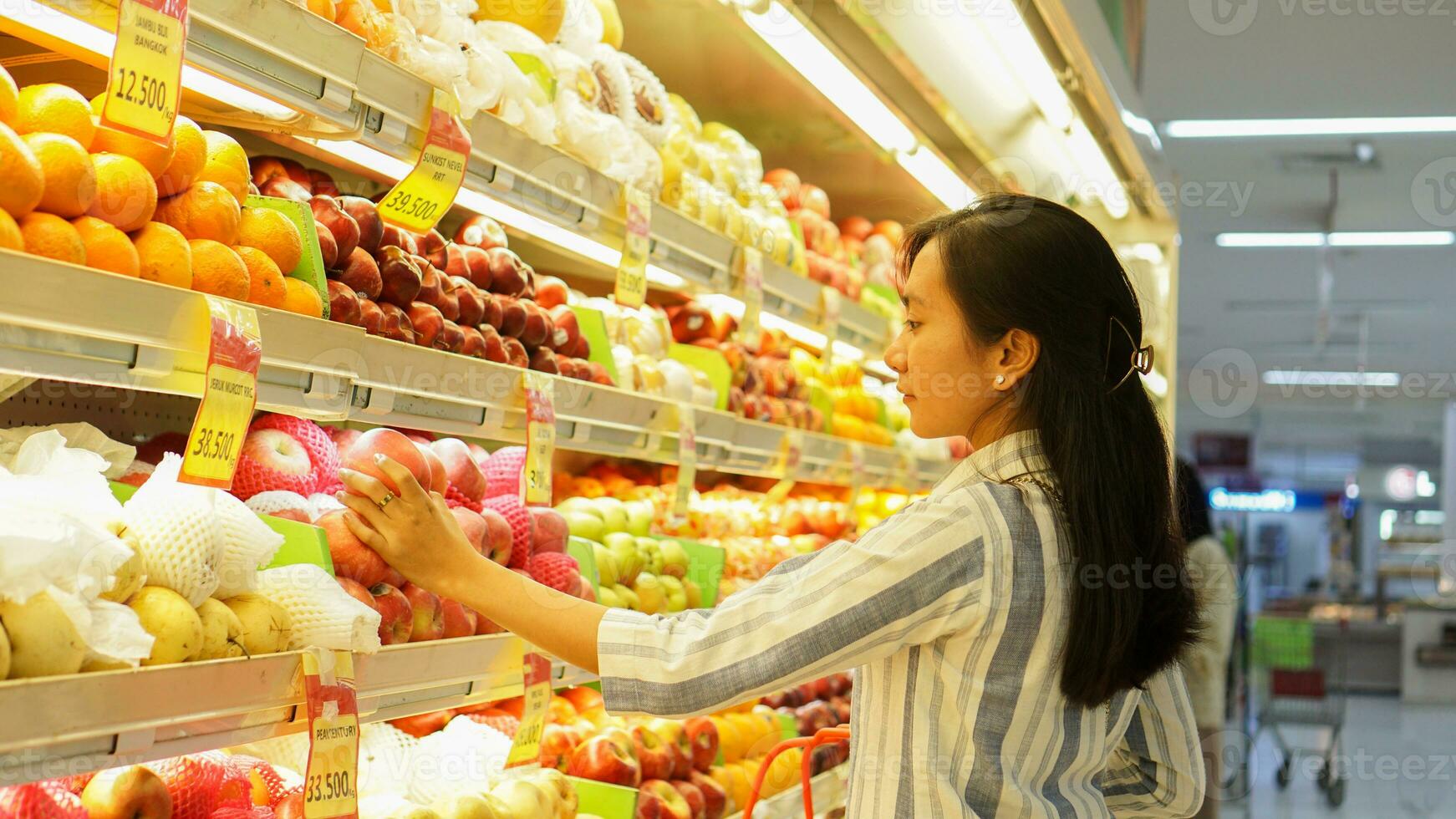 portrait of a beautiful Asian or Indonesian woman buying fruit arranged on fruit rack in a supermarket photo