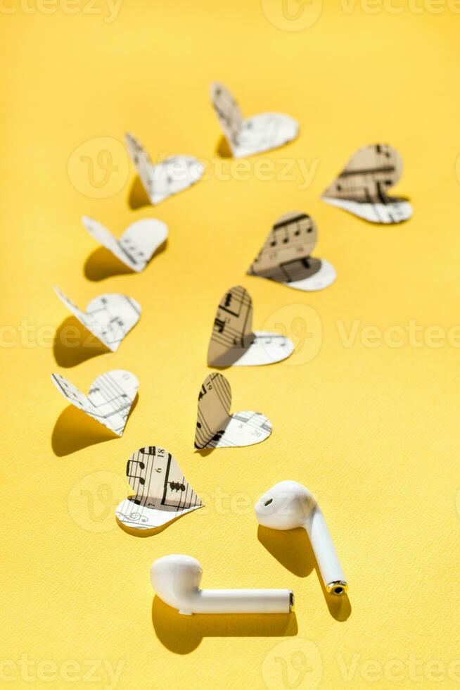 Wireless headphones and hearts cut from the music text on a yellow background. Love to listen to music using gadgets. Vertical view photo