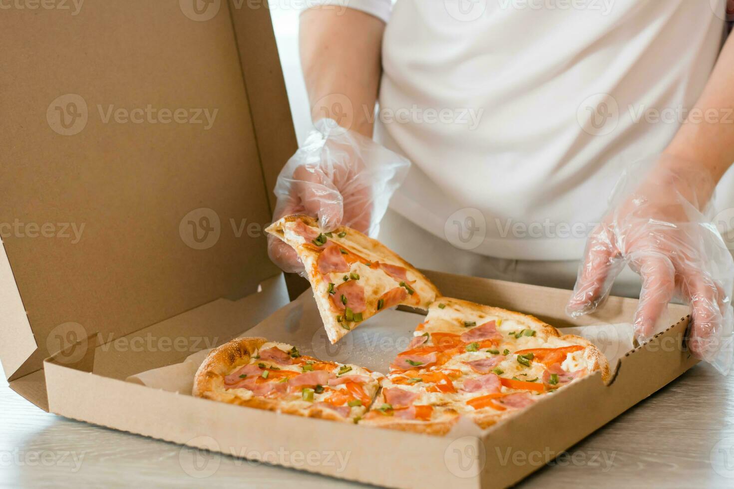 Takeaway  food. A woman in disposable gloves takes a slice of pizza from a cardboard box on the table in the kitchen. photo
