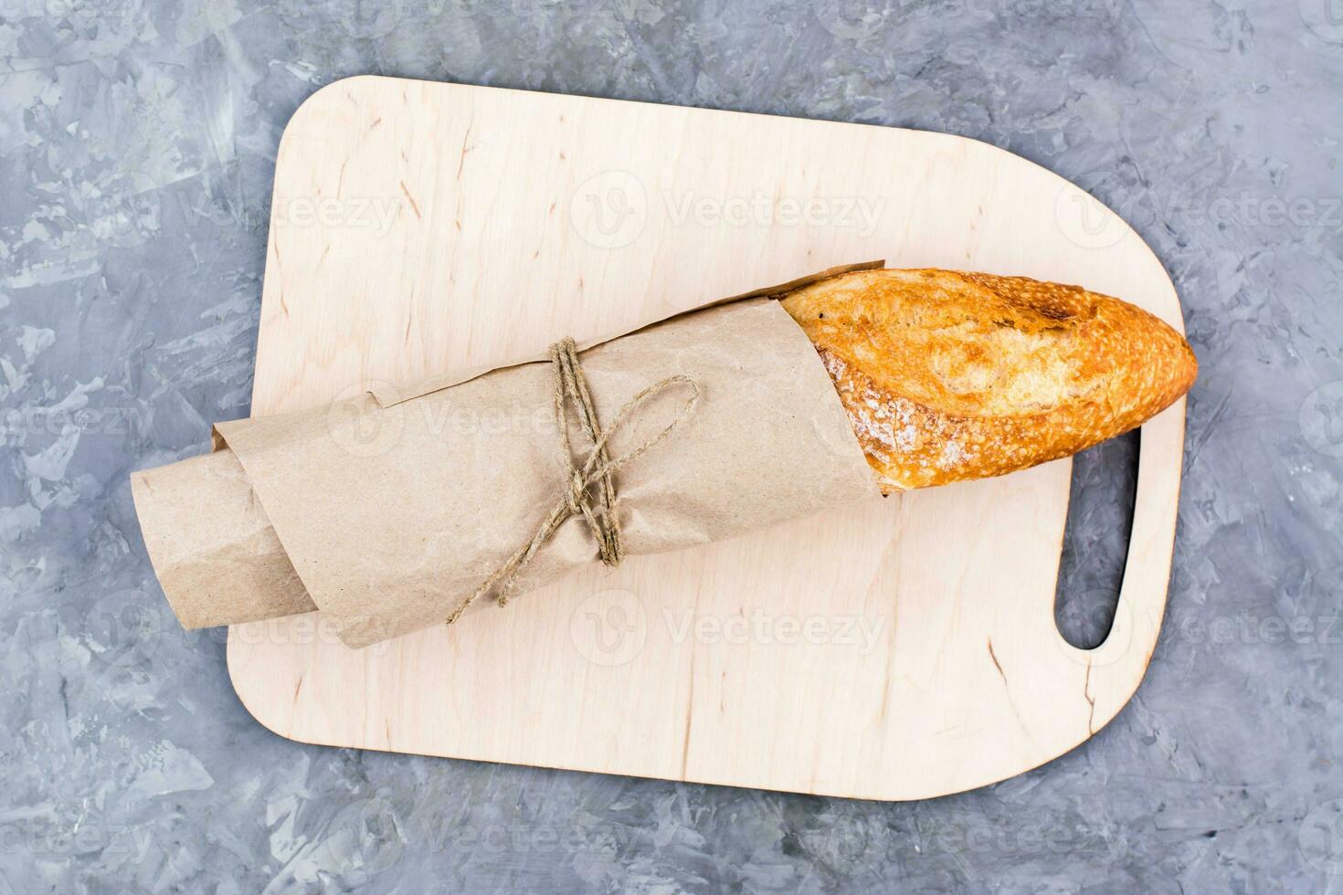Fresh baguette in wrapping paper on cutting board on table. Top view photo