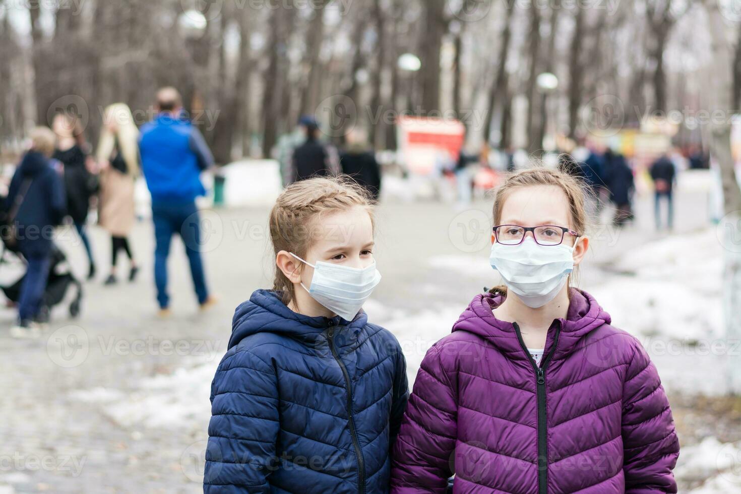 Two girls in protective face masks in the park, at a distance from the crowd of people. Walk the new normal photo