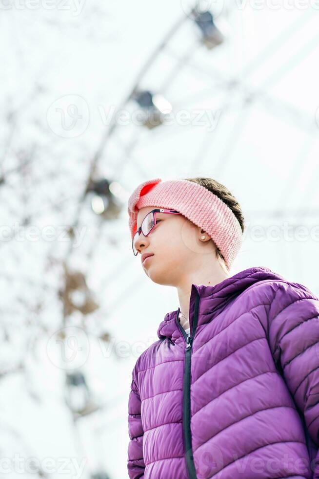 Serious girl in glasses in early spring in a city amusement park against the background of a ferris wheel photo