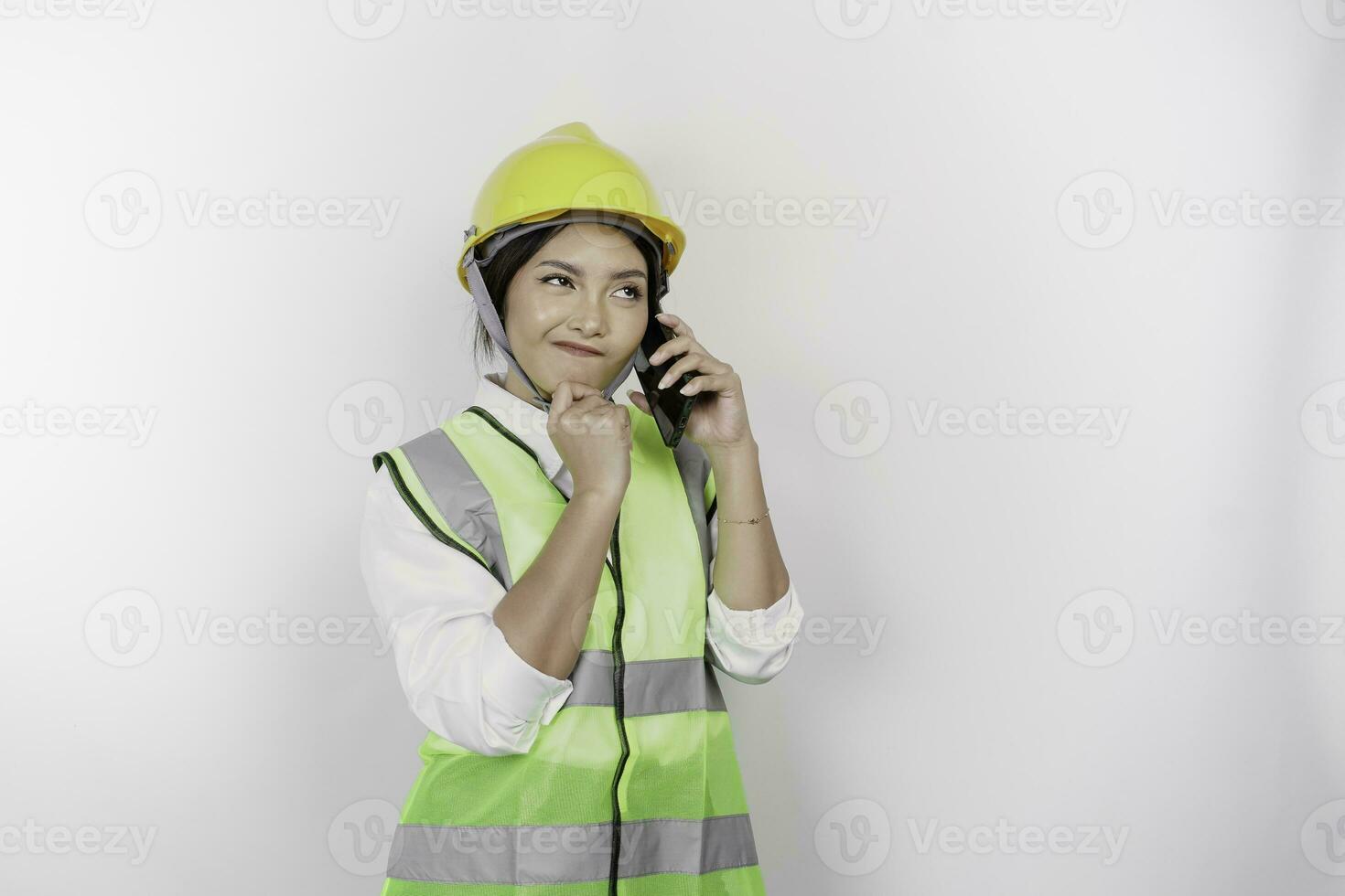 A thoughtful young woman labor worker wearing safety helmet and vest while getting a phone call and hand on her chin, isolated by white background. Labor's day concept. photo