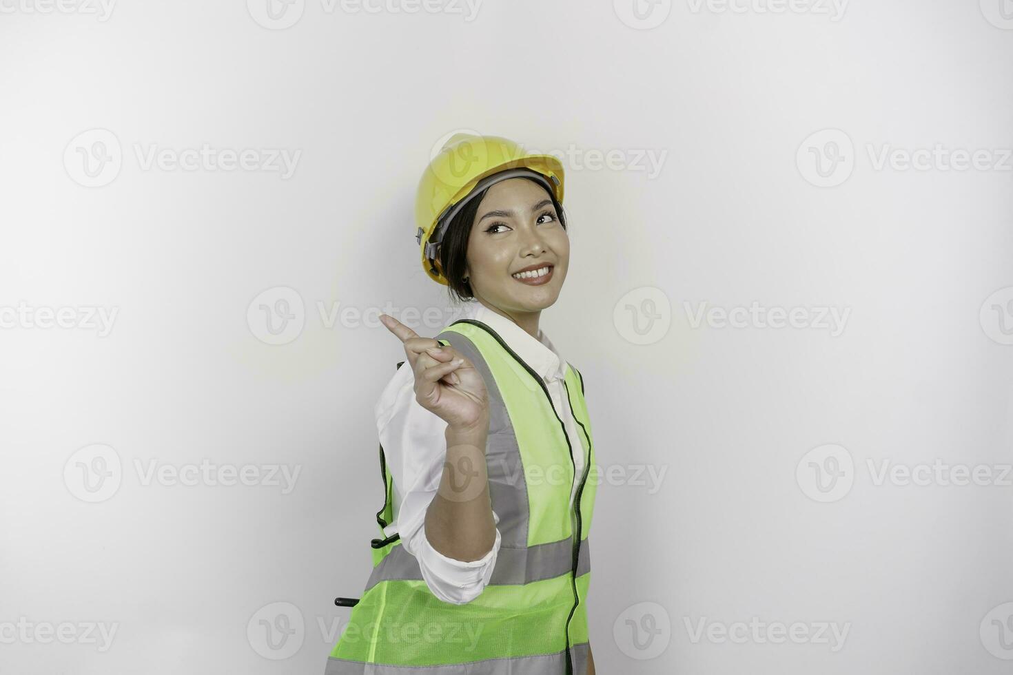 A smiling Asian woman labor wearing safety helmet and vest, pointing to copy space beside her, isolated by white background. Labor's day concept. photo