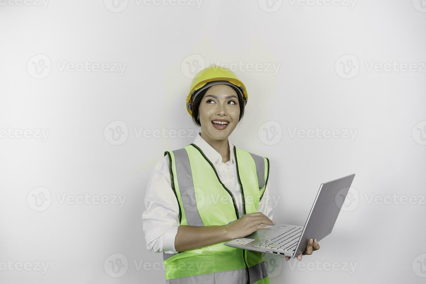 A thoughtful young woman labor worker wearing safety helmet and vest while holding her laptop and looking aside to copy space, isolated by white background. Labor's day concept. photo