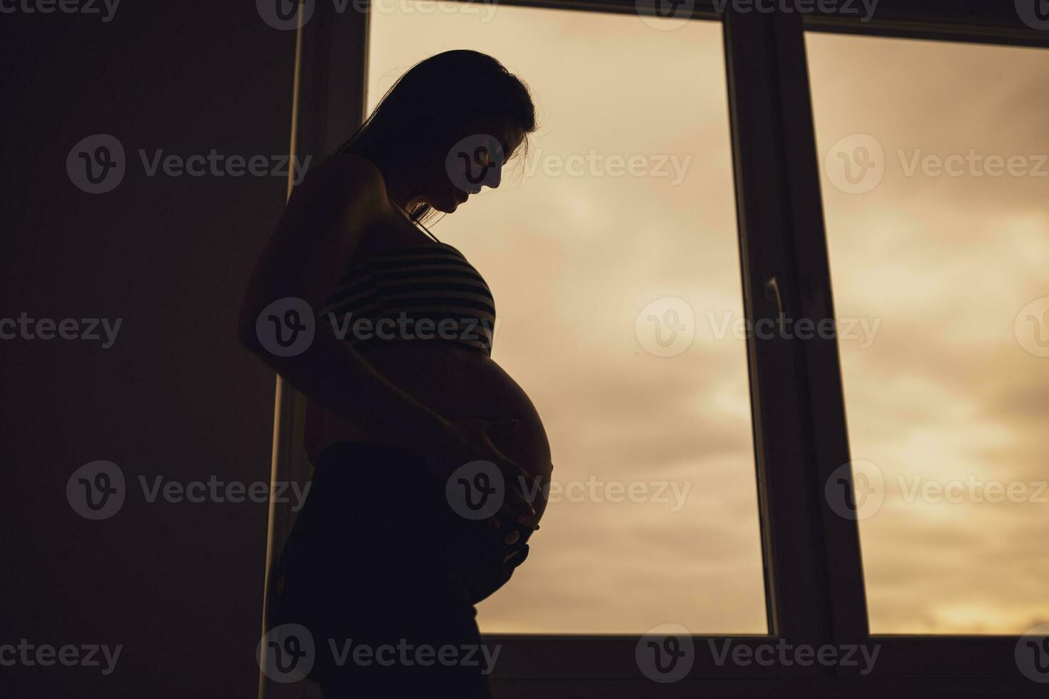 Pregnant woman standing beside window and holding her belly. Silhouette of pregnant woman. photo