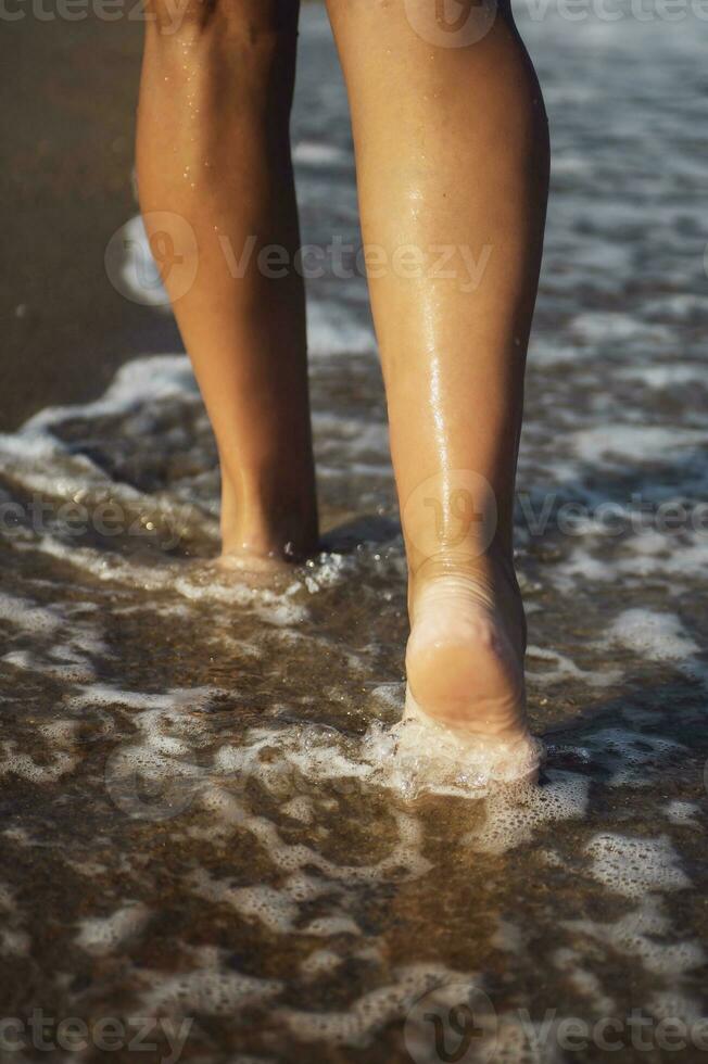 de cerca detalle de hembra piernas caminando a lo largo el costa. De las mujeres piernas y pies caminar a lo largo el arena y mar ondas, a lo largo el playa. relajación viaje concepto. vertical foto. foto