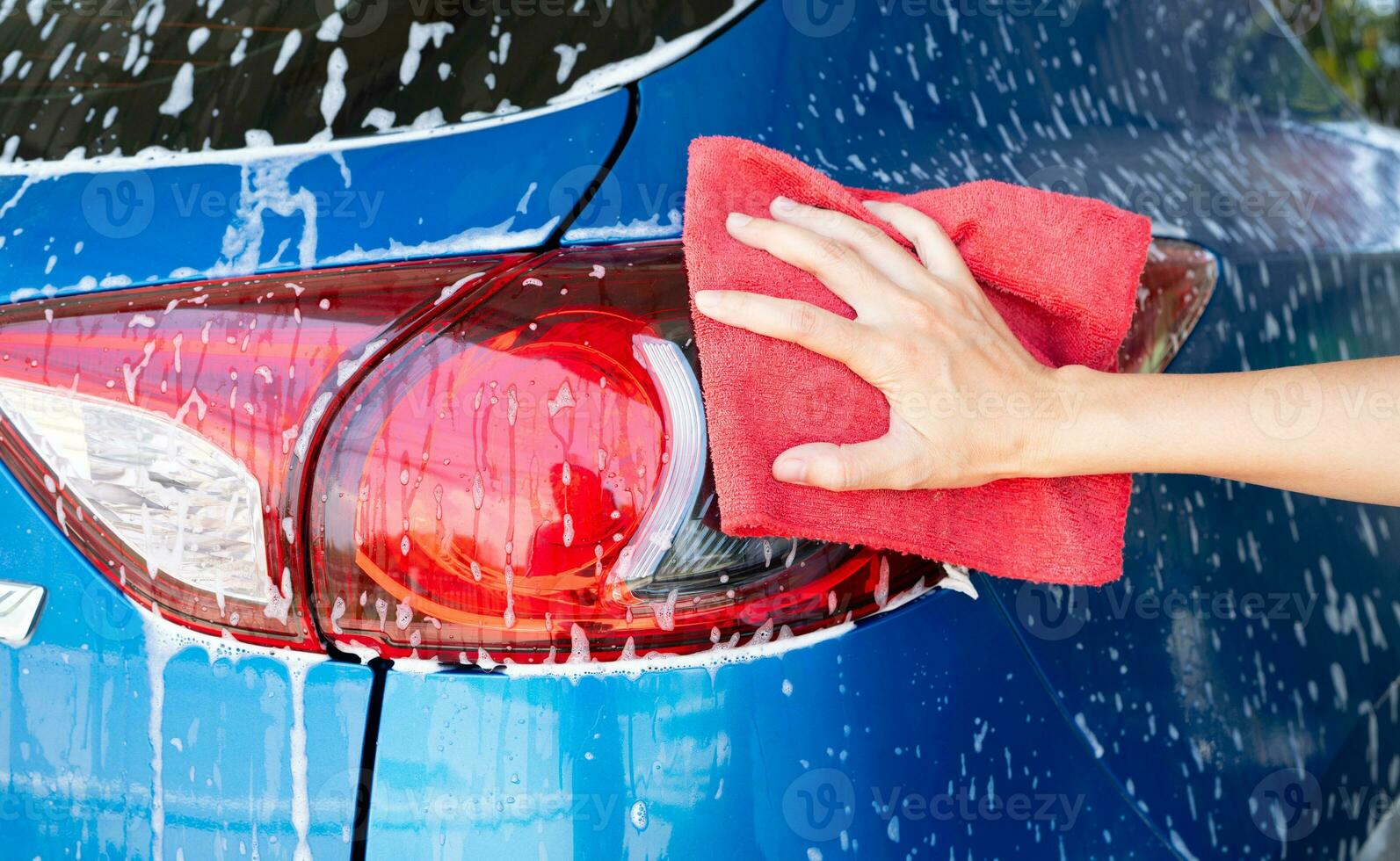 Car wash service. Car covered with white soap foam. Man hand holding red microfiber cloth and polish tail light of blue car. Auto care service business concept. Man cleaning and detailing luxury car. photo