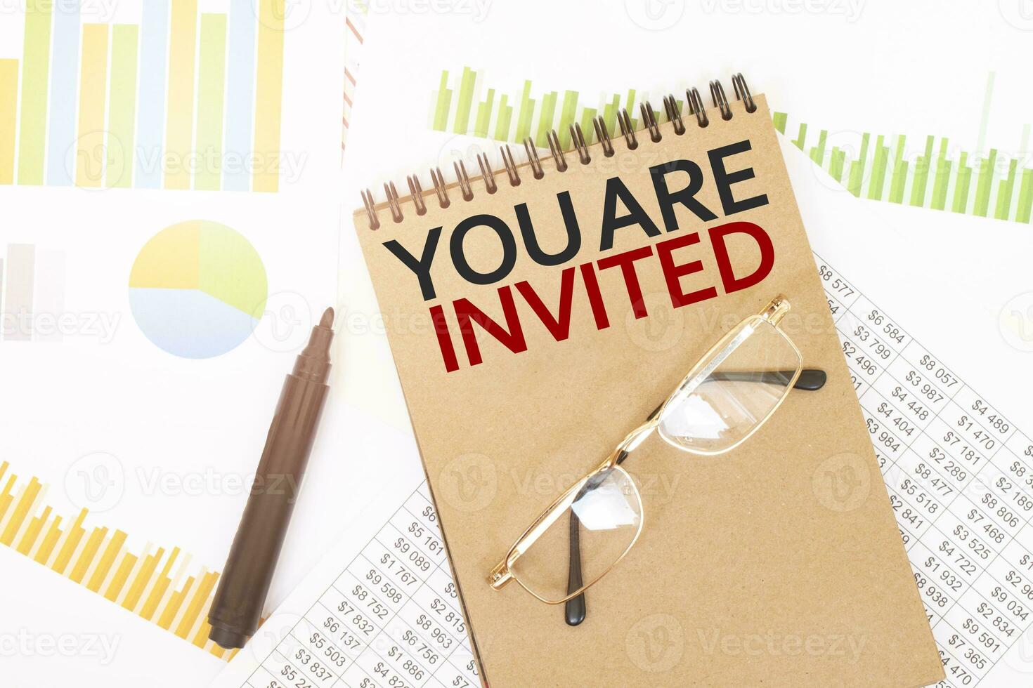 In a craft colour notebook is a YOU ARE INVITED inscription, next to pencils, glasses, graphs and diagrams. photo