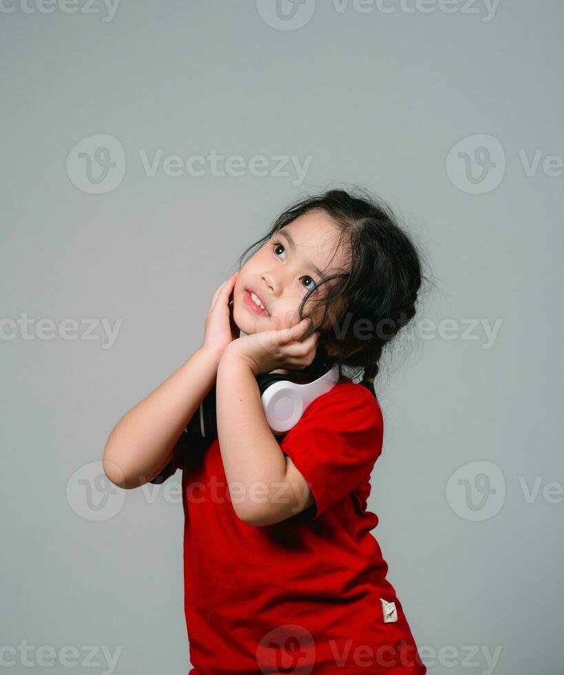 Cheerful happy asian children girl with black hair listening to music songs in headphones on a gray background. Portrait cute girl waring red shirt and looking at camera studio lighting. photo