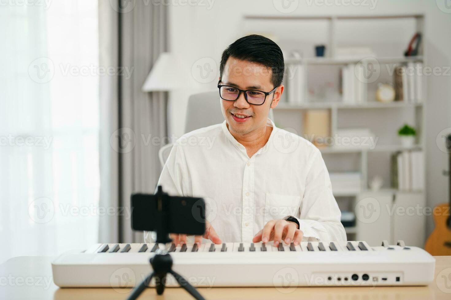 Asian man wearing white headphones play piano and sing a song and learning online with mobile phone and compose writing song or record sound with a microphone and use computer recording music program photo