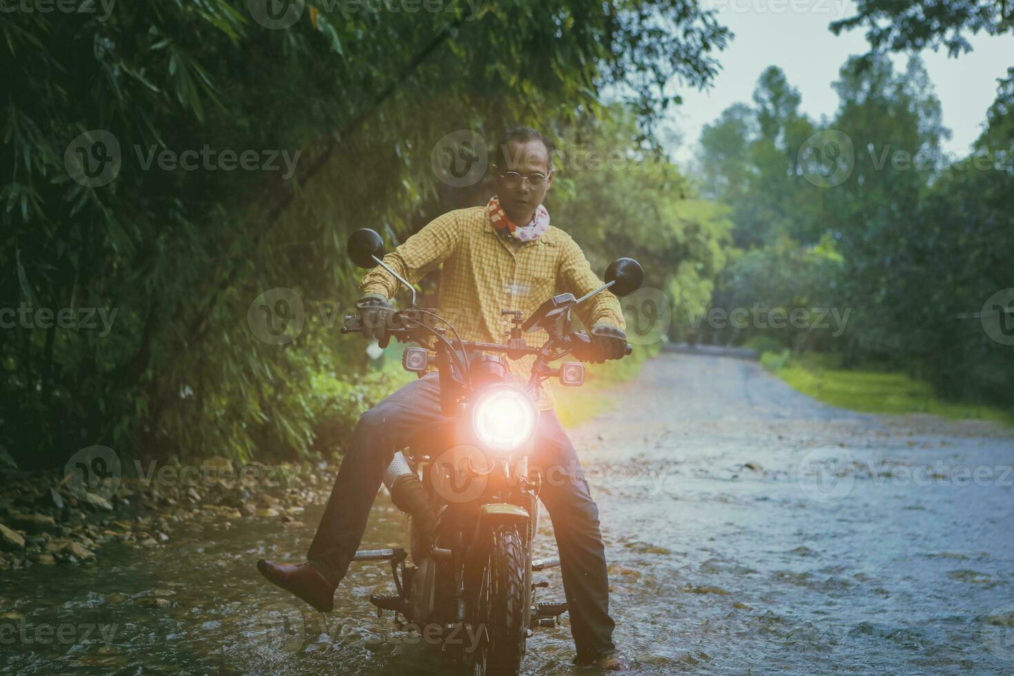 asian man riding small enduro motorcycle crossing flowing creek in forest among rain falling photo