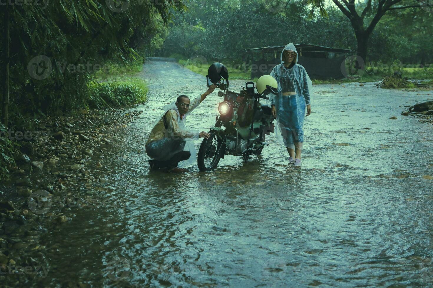 couples of asian biker wearing plastic rain clothes  maintenance small enduro motorcycle in shallow creek among rain falling photo