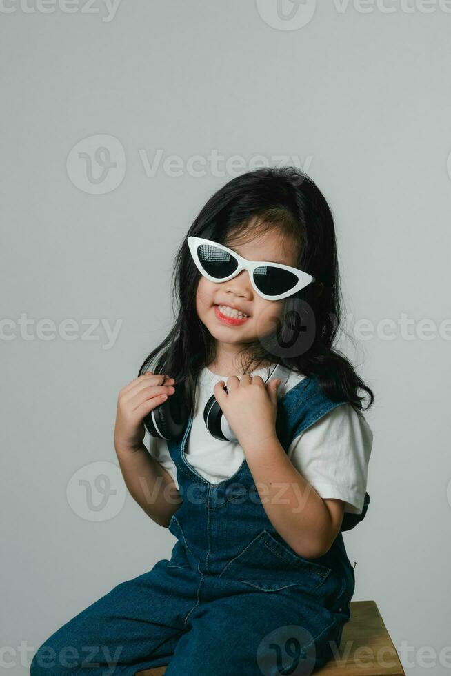 Portrait of relaxed cute little asian girl child funny smiling and laughing wearing white headphones and glasses, keeps hands on her ears, posing and smiling enjoying isolated over gray background. photo