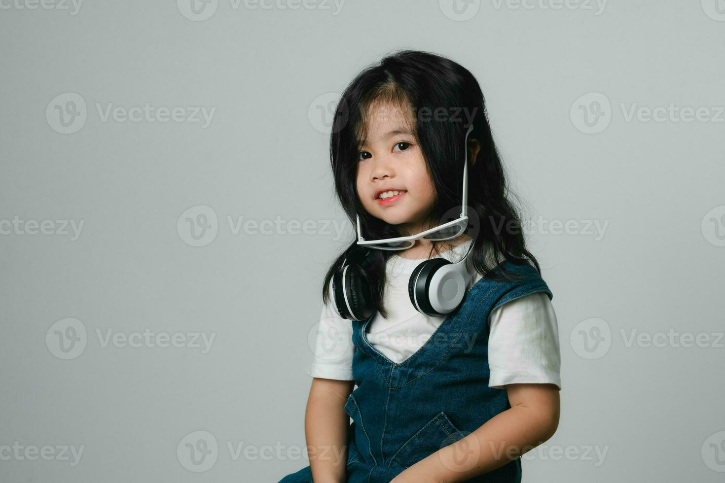 Portrait of relaxed cute little asian girl child funny smiling and laughing wearing white headphones and glasses, keeps hands on her ears, posing and smiling enjoying isolated over gray background. photo