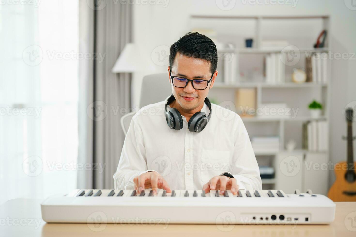 Asian man wearing white headphones play piano and sing a song and learning online with mobile phone and compose writing song or record sound with a microphone and use computer recording music program photo