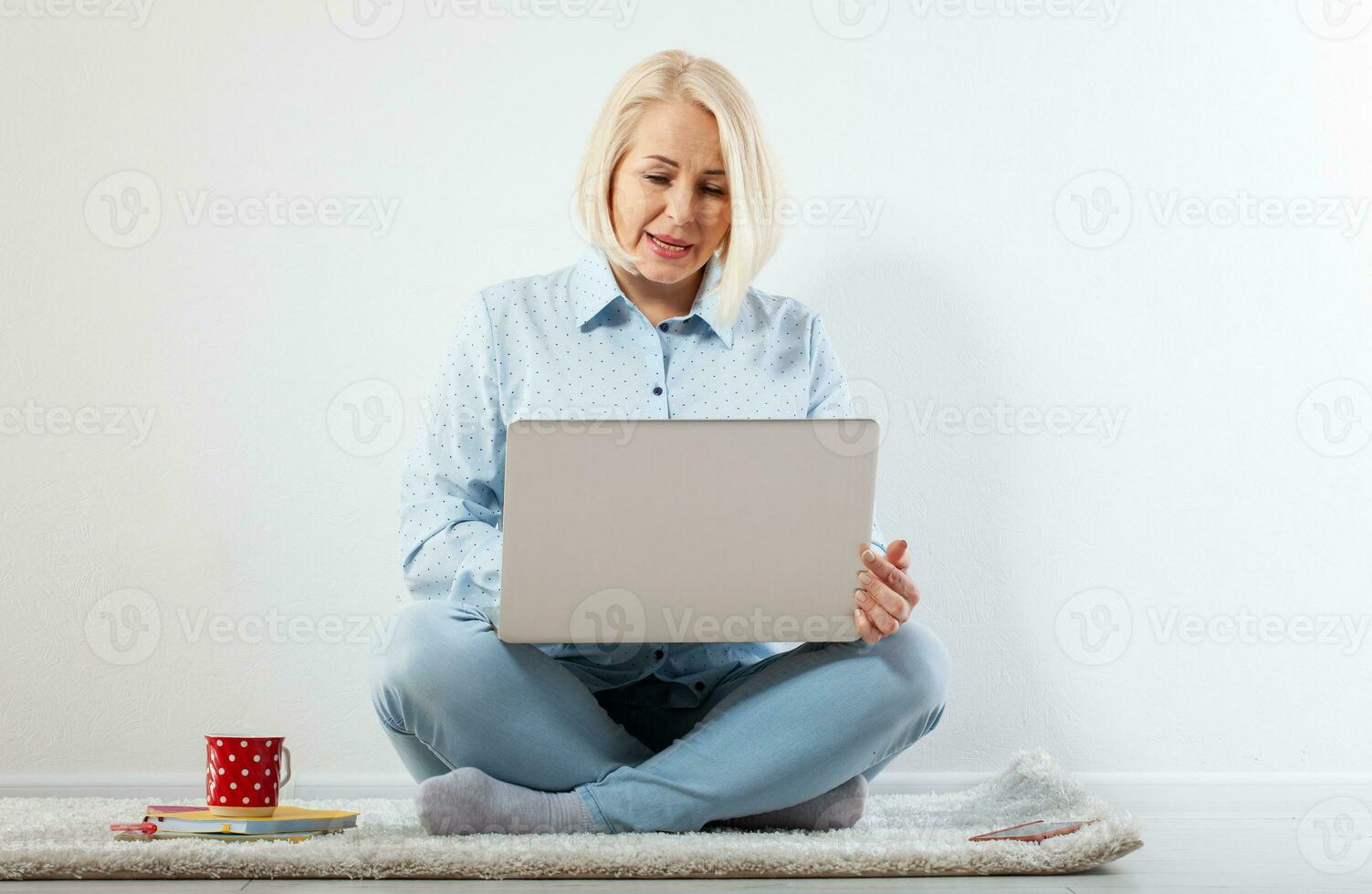 Happy middle aged woman sitting relaxed on the floor using laptop for entertainment. The concept of leisure and work with a cup of coffee at home. photo