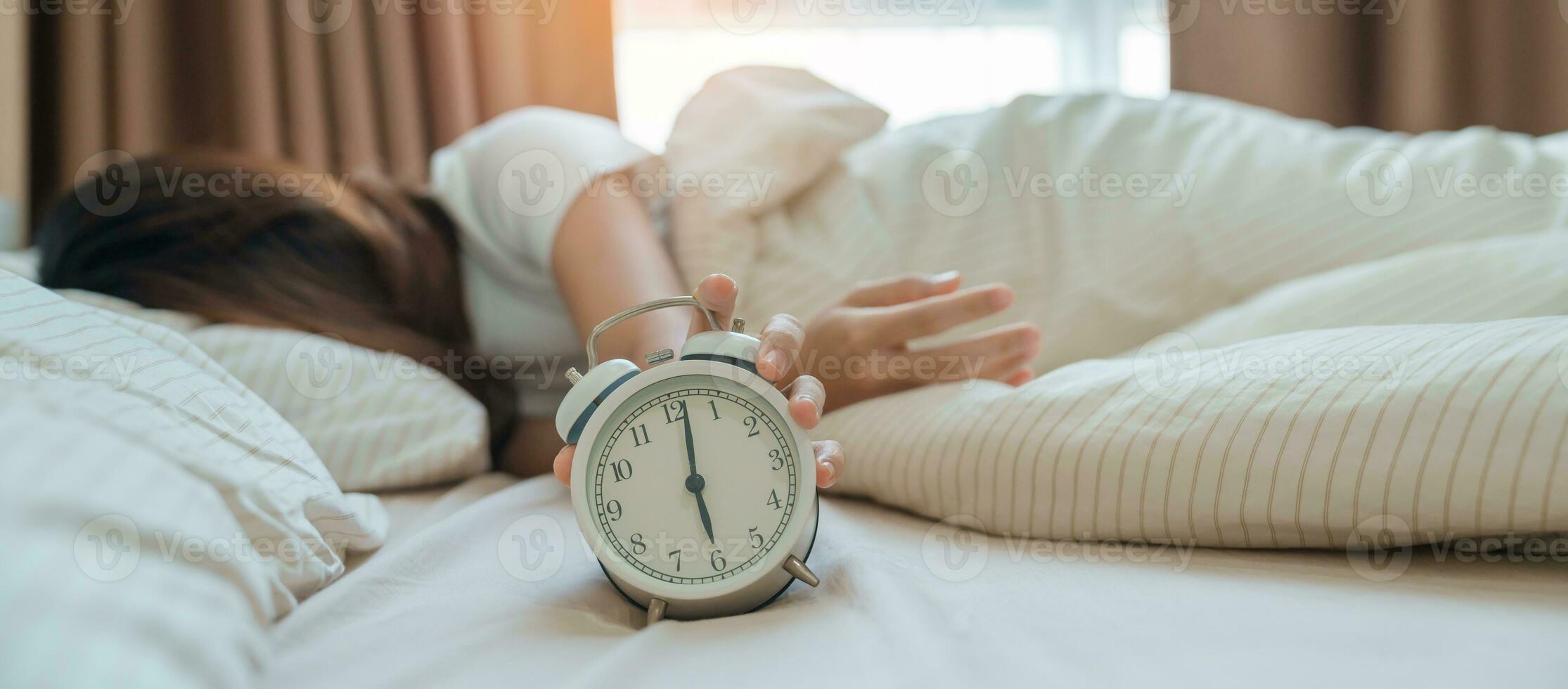 alarma reloj y asiático mujer mano detener hora en cama mientras durmiendo, joven adulto hembra despertar arriba tarde en el temprano Mañana. relajante , somnoliento, diario rutina y tener un bonito día conceptos foto